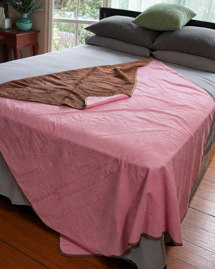 Pink side of the waterproof blanket draped over a bed with Coffee reverse visible
