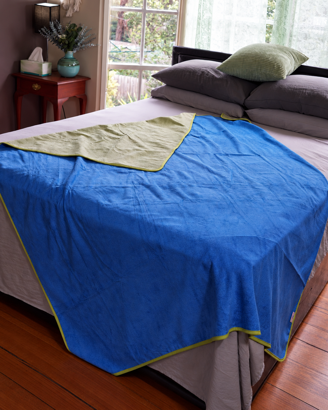 Capri Blue side of the waterproof blanket draped over a bed with Matcha Mist reverse visible
